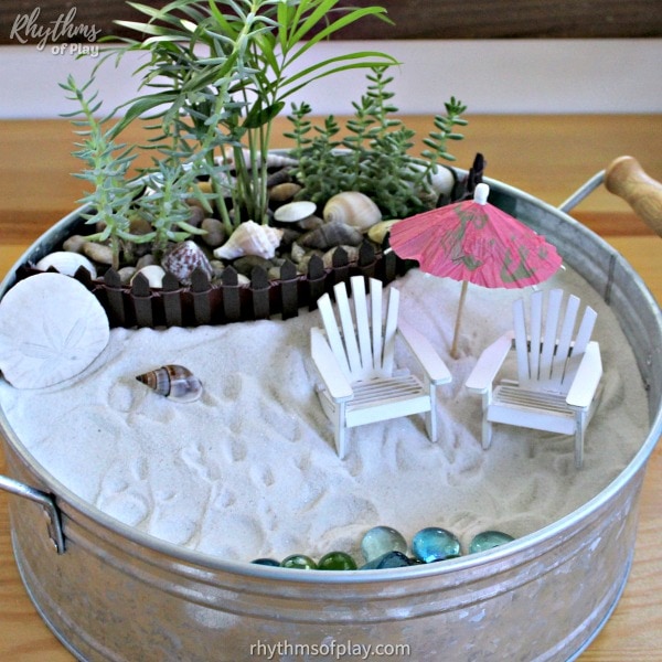 beach-themed mini garden with white adirondack chairs, pink umbrella, sea shells, glass stones, wooden fence and plants inside a metal bucket